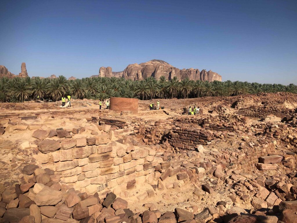 Archaeologists working among ancient stone walls and archaeological remains in AlUla, Saudi Arabia, with palm groves and sandstone mountains in the background.