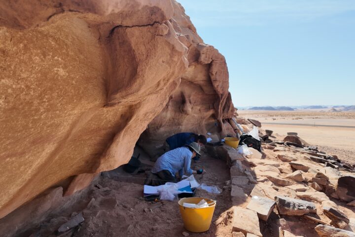 Archaeologists working beneath a sandstone rock shelter in Saudi Arabia's desert landscape.