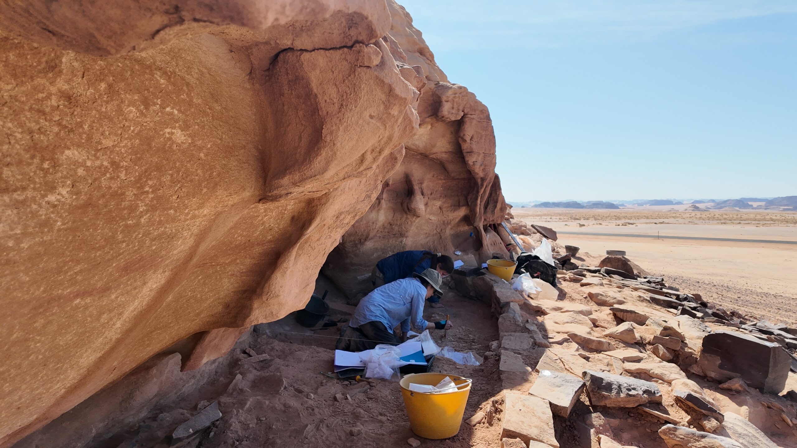 Archaeologists working beneath a sandstone rock shelter in Saudi Arabia's desert landscape.