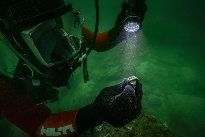 Underwater archaeologist holding an ancient bronze ring depicting the Lighthouse of Alexandria while examining it with a flashlight.
