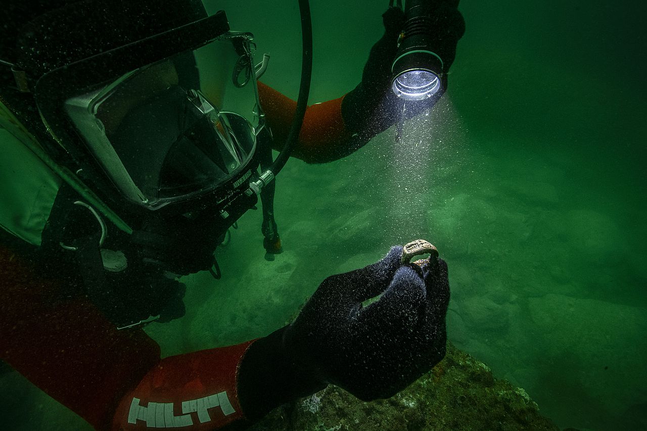 Underwater archaeologist holding an ancient bronze ring depicting the Lighthouse of Alexandria while examining it with a flashlight.